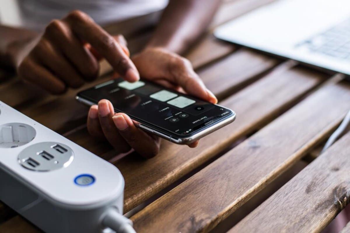 Person using a smartphone next to a laptop and smart power strip on a wooden table.