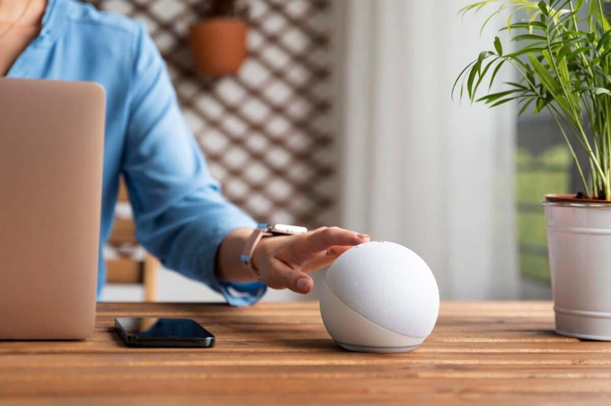 Person using a smart speaker on a wooden desk, alongside a laptop and smartphone, in a light-filled room with plants.