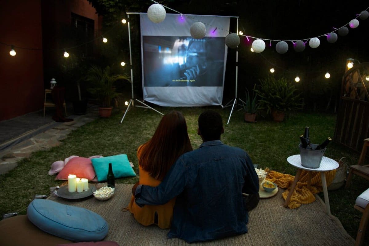 A couple enjoys an outdoor movie night in a cozy backyard setting with snacks and string lights.