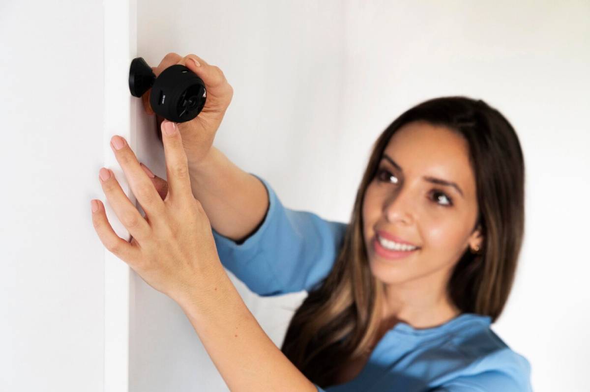 A person in a blue shirt mounts a round black device on a white wall, using both hands for precision.