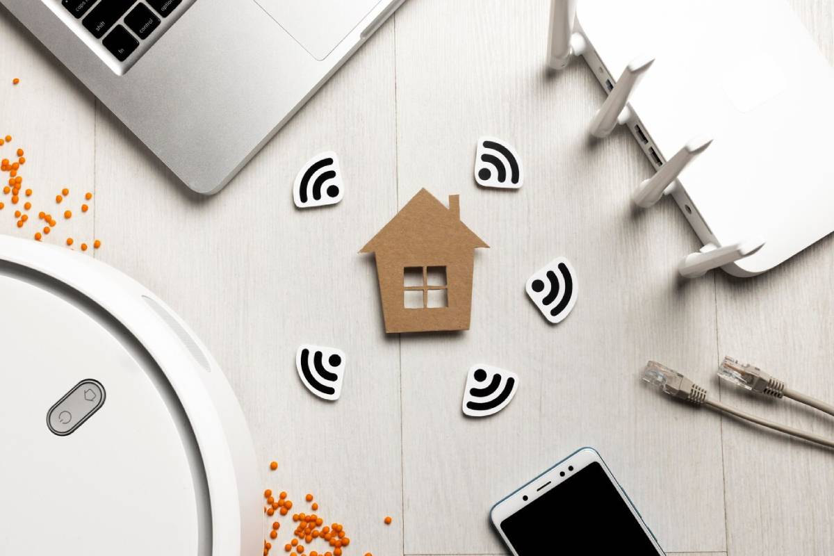 A flat lay of smart home devices: a laptop, smartphone, router, and a cardboard house surrounded by Wi-Fi symbols and orange beads.