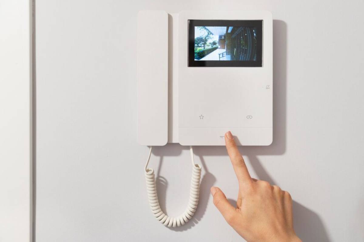 A hand hovers over a smart doorbell mounted on a wall, showing an outdoor view on its screen.