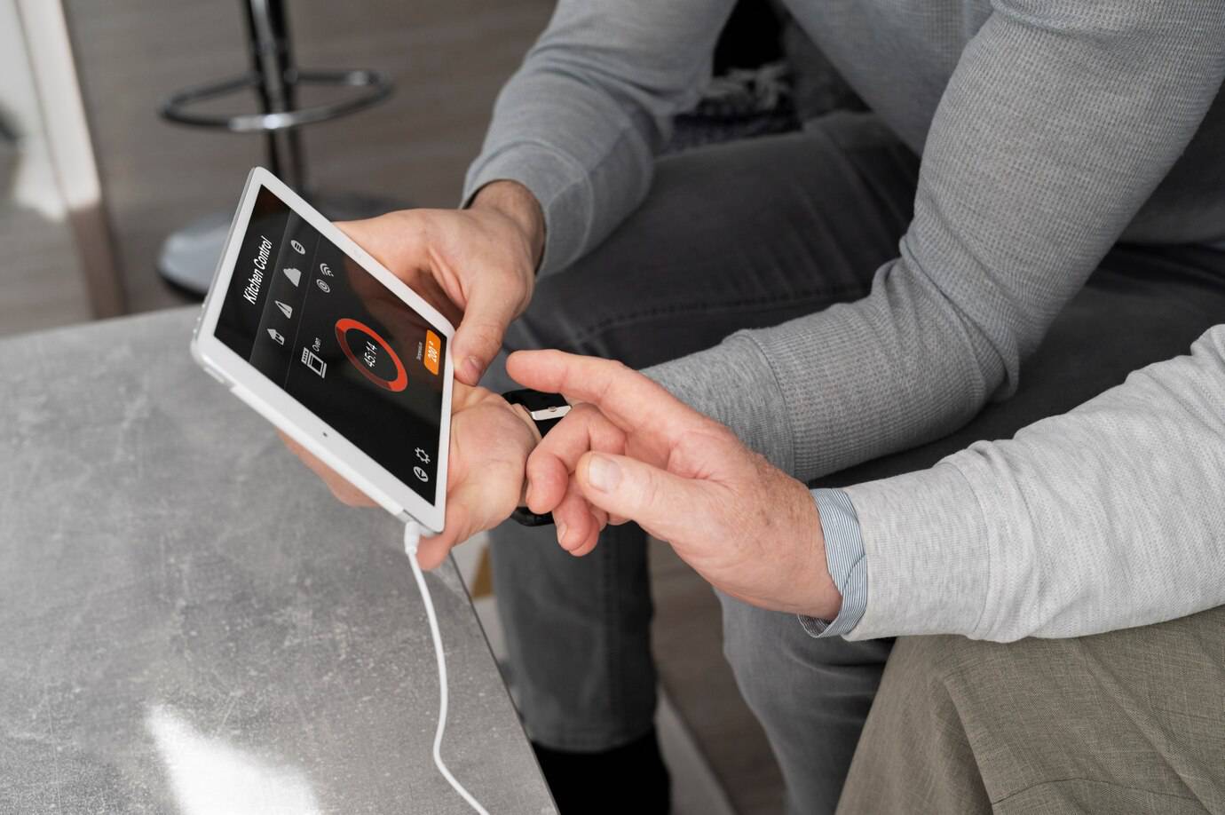 Two people interact with a tablet displaying a kitchen control app, while connected by a white charging cable. One points at the screen.