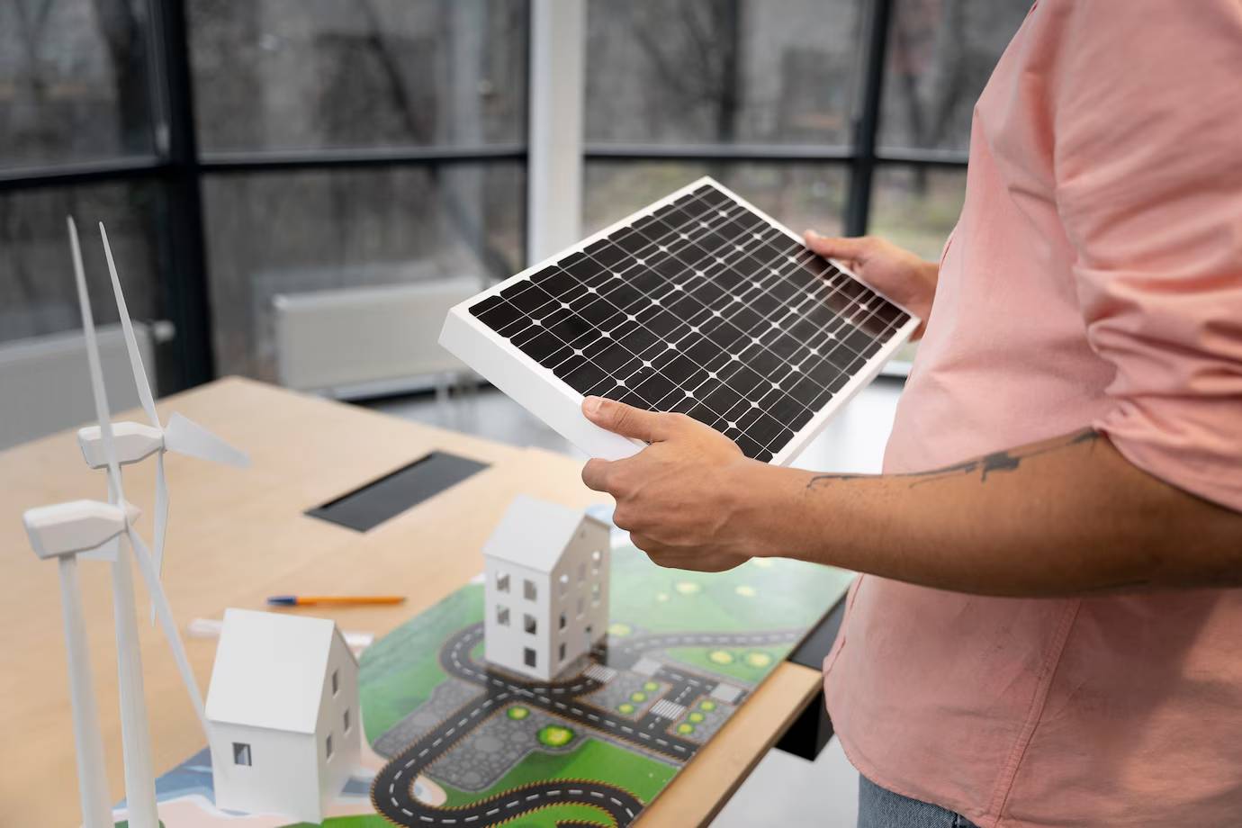 A person holds a solar panel above a model landscape with wind turbines and houses, emphasizing sustainable energy design.
