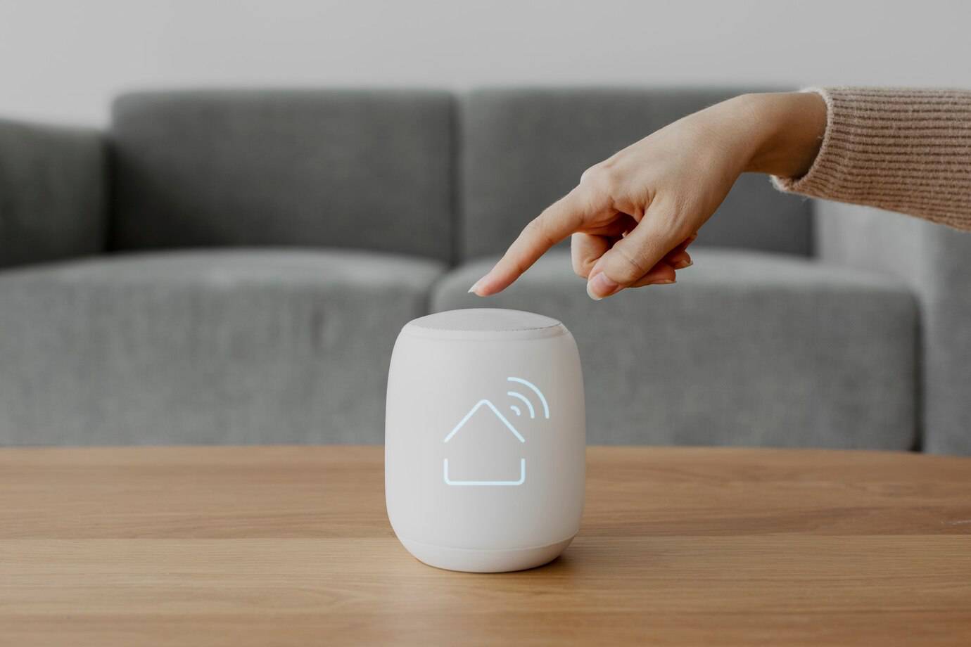 A hand presses a smart home device on a wooden table, featuring a glowing house icon, with a gray couch blurred in the background.