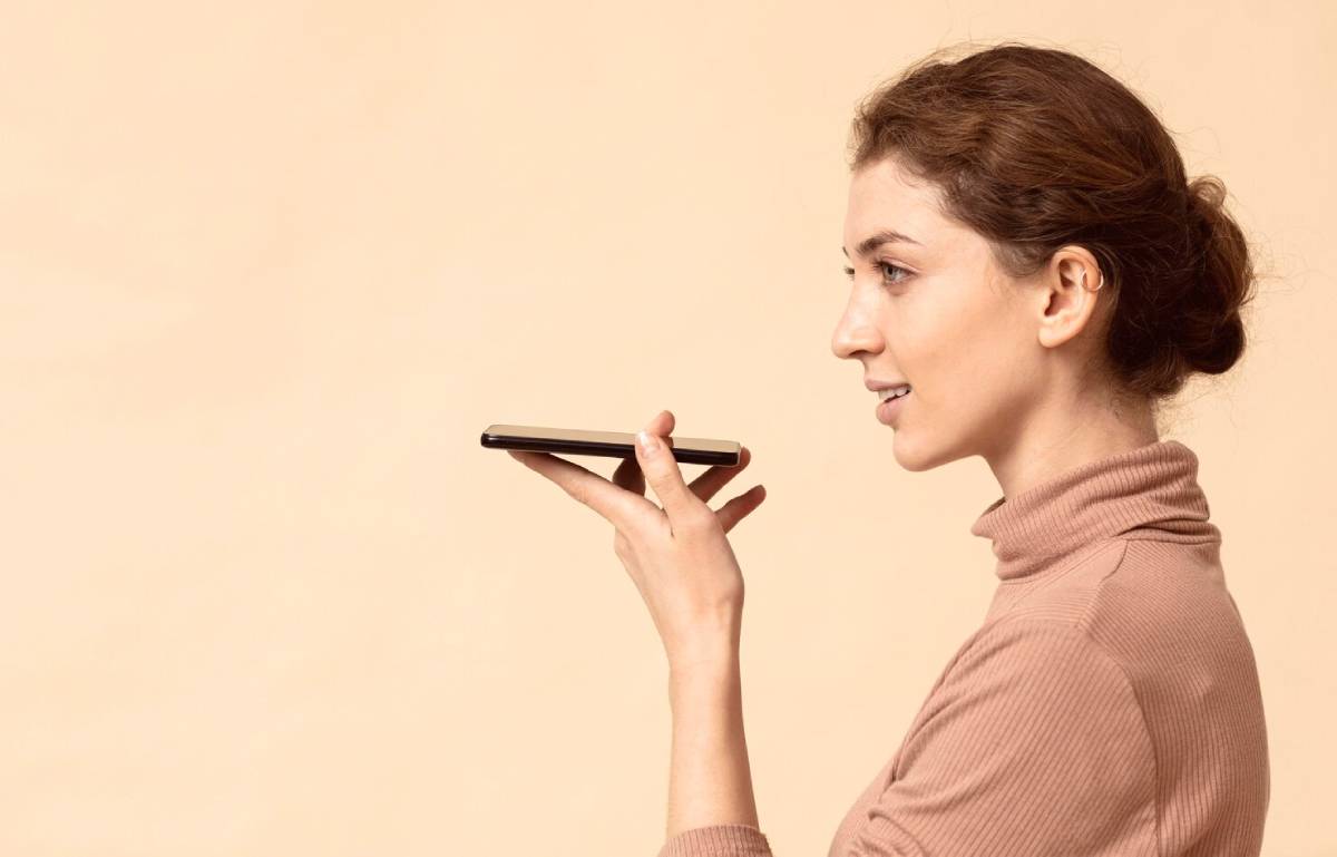 A woman holds a smartphone to her mouth, wearing a cozy, light pink turtleneck sweater against a soft beige background.