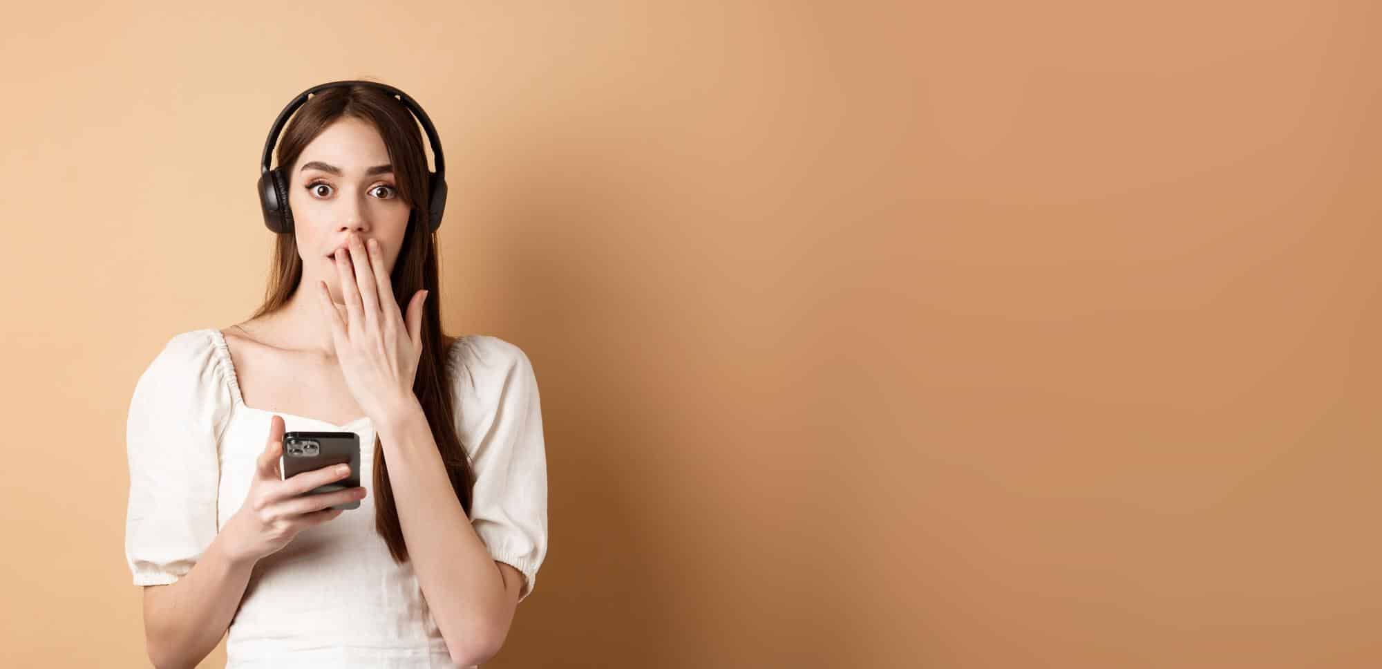 A person wearing headphones holds a smartphone, appearing contemplative in front of a warm, beige background.