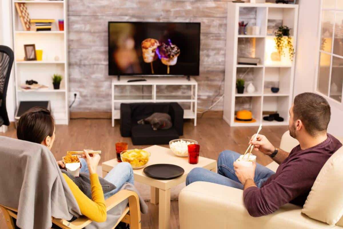 A man and woman enjoy takeout while watching TV at home, with snacks and a sleeping cat nearby. Cozy living room setting.