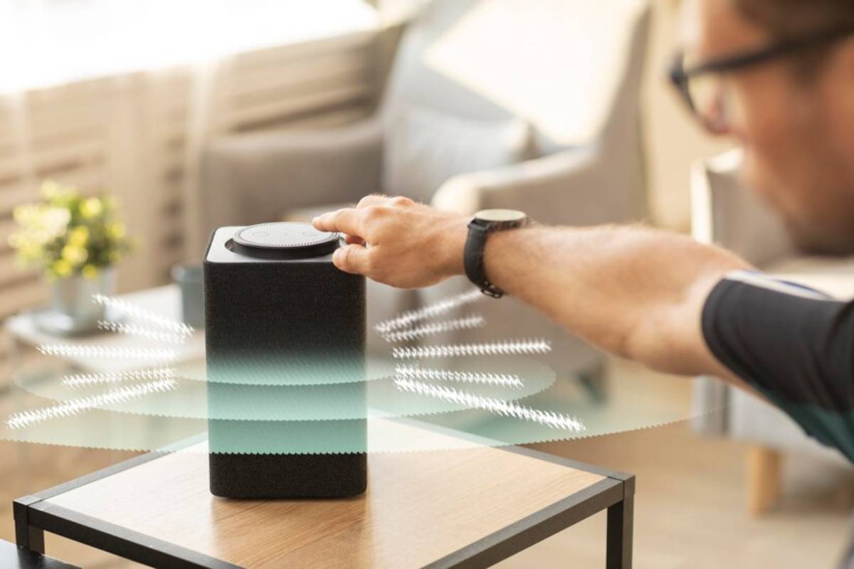 A person’s hand reaches toward a modern speaker on a wooden table, with sound waves visually emanating from the device in a bright room.