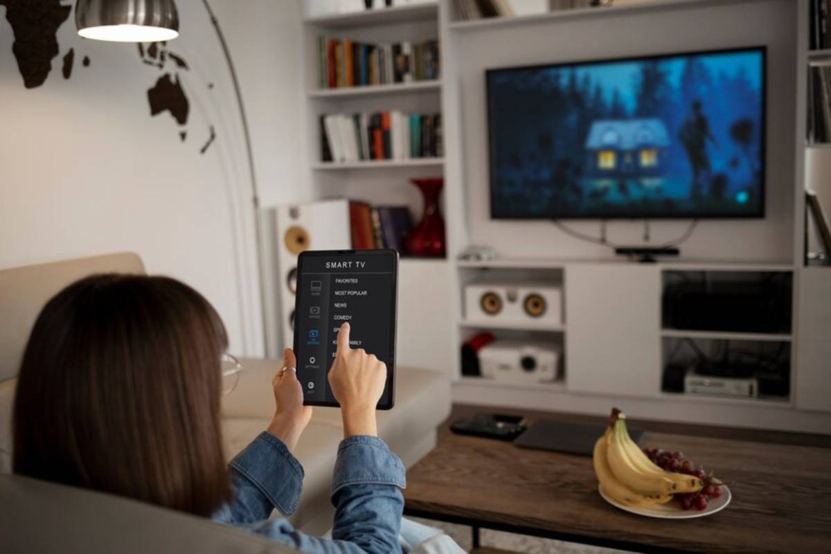 A person controls a smart TV with a tablet, displaying a cozy scene on-screen, with snacks on the table in a stylish living room.