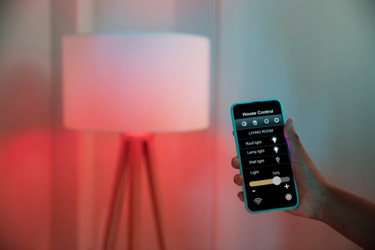 A hand holds a smartphone displaying smart home controls, with a lamp emitting a soft red glow in the background.