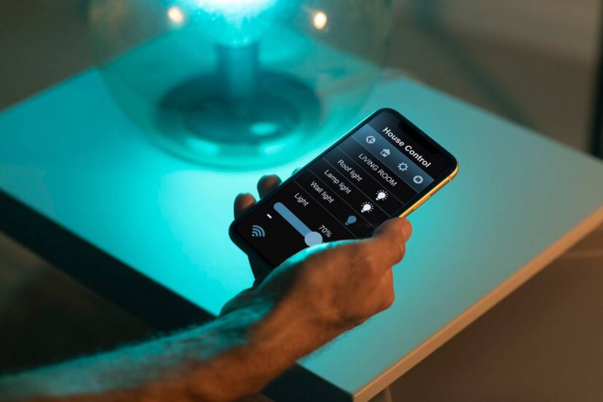 A hand holding a smartphone displaying a smart home app, controlling lighting settings, next to a glowing lamp.