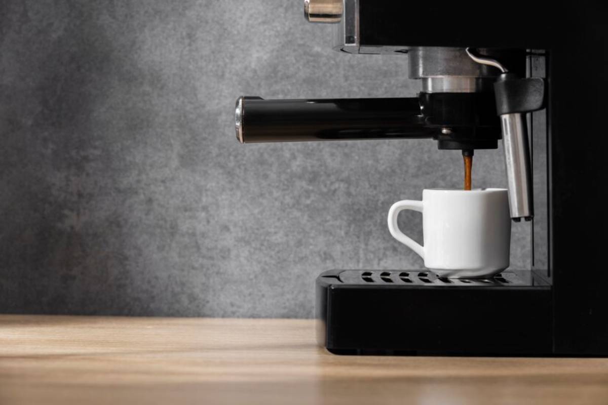 A black coffee machine pours espresso into a small white cup on a wooden countertop against a textured gray background.