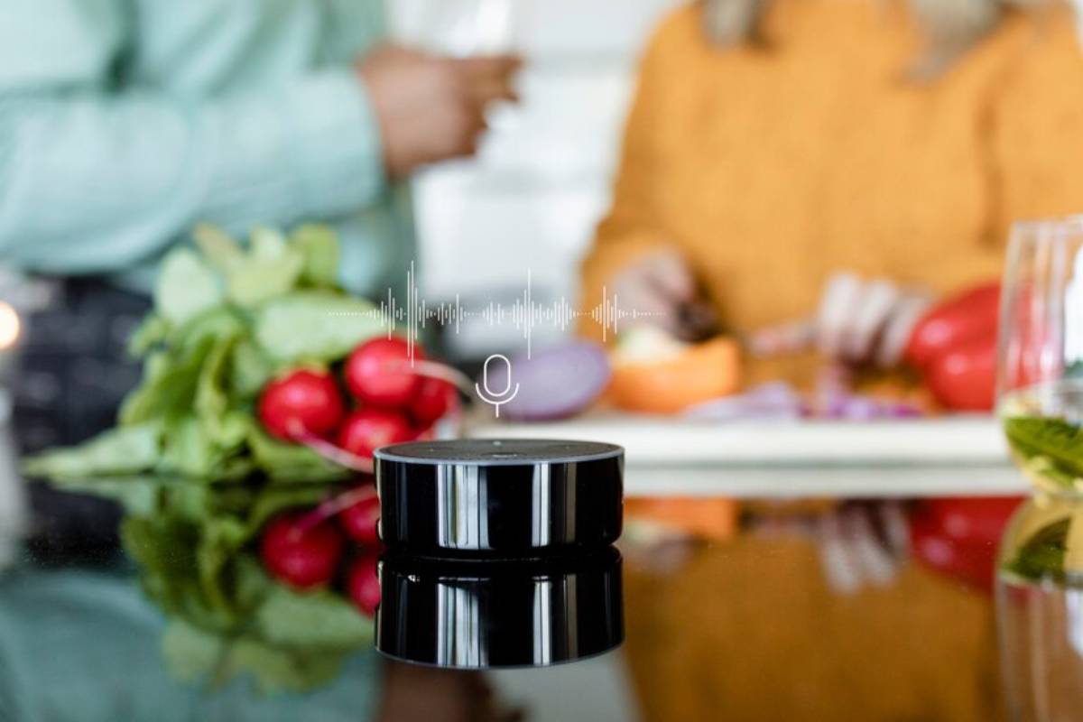 A sleek black smart speaker sits on a shiny surface, surrounded by fresh vegetables, with people preparing food in the background.