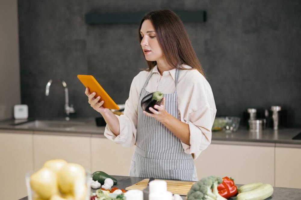A woman in an apron holds an eggplant and a tablet in a modern kitchen with various vegetables on the counter.