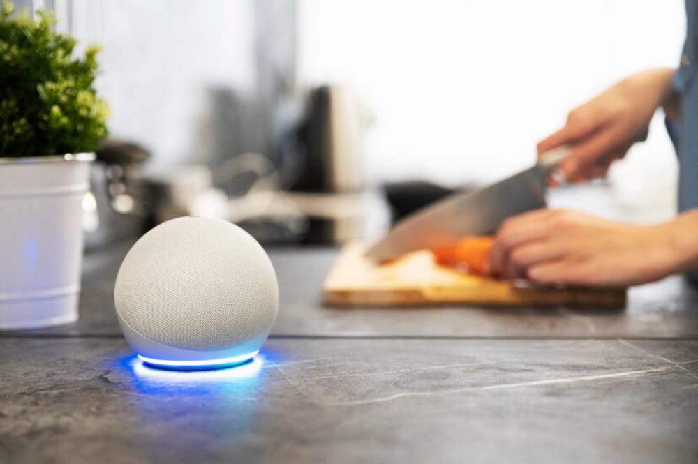 A white smart speaker glows blue on a kitchen counter, while a person chops carrots on a wooden cutting board in the background.