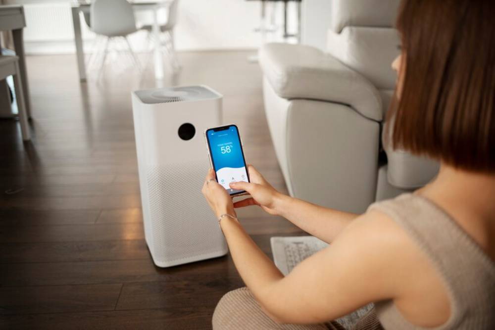 A person sits on the floor, checking air quality data on their smartphone, while a white air purifier stands nearby in a modern living space.