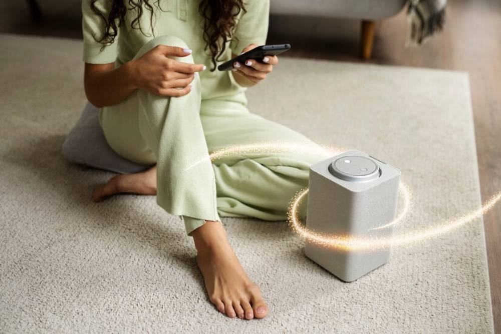 A woman in a pale green outfit sits on a carpet, using her phone beside an air purifier.