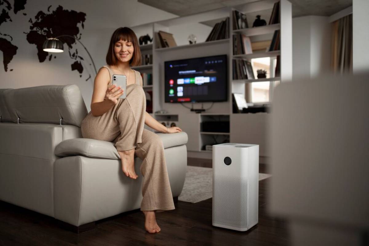 A person sits on a light gray sofa in a modern living room, holding a smartphone beside a sleek white air purifier.
