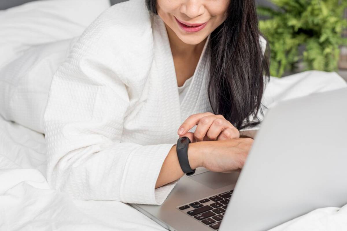 A person in a white robe lies on a bed, using a laptop, while wearing a sleep tracker on their wrist. Green plants are visible in the background.