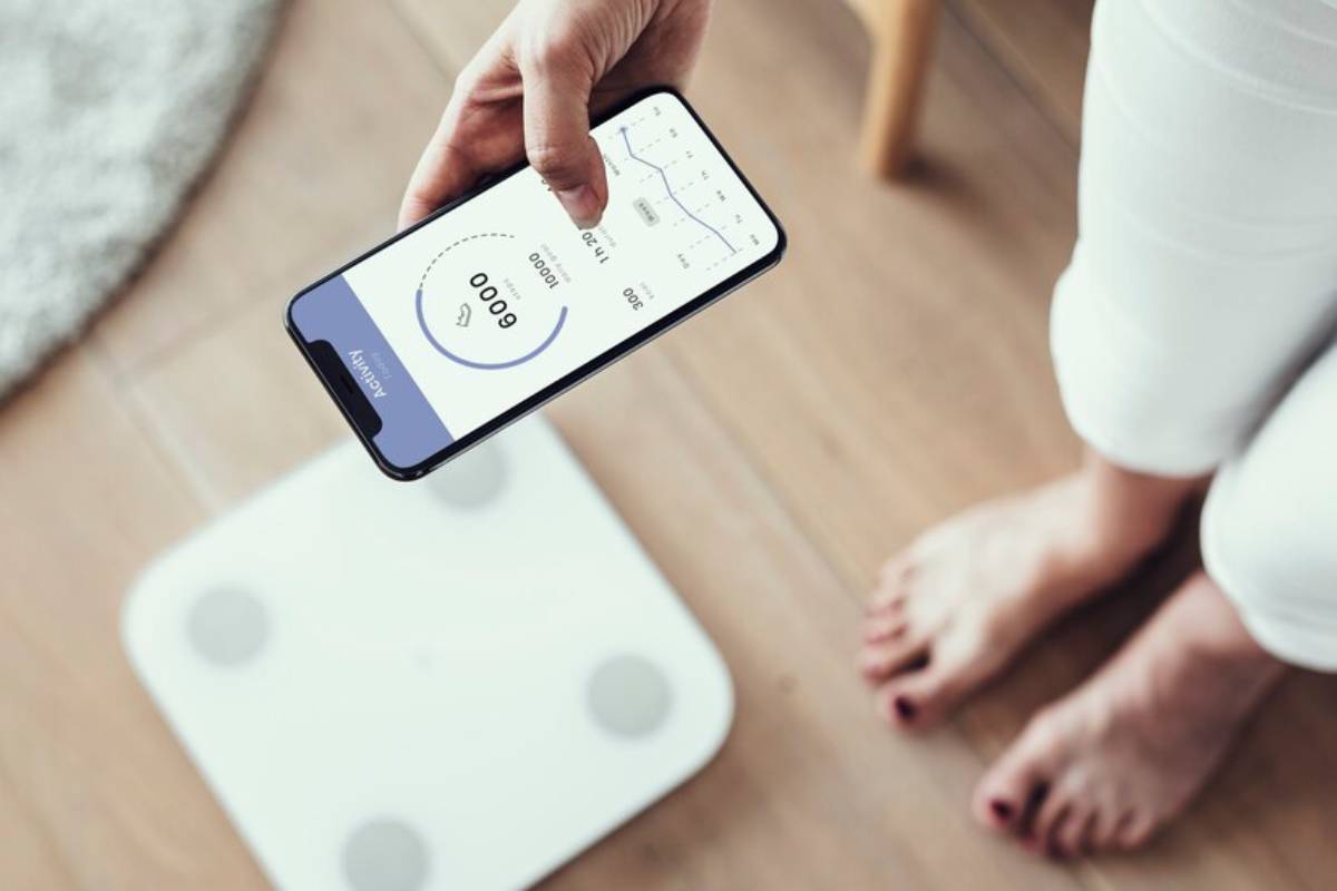 A person in white pants checks a fitness app on their smartphone while standing next to a digital scale on wooden flooring.