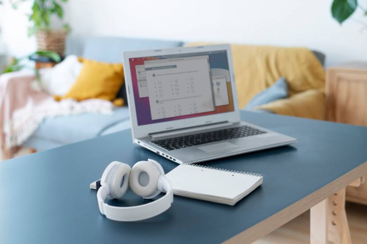 A laptop displaying a software interface sits on a desk beside white headphones and a notepad, with a cozy sofa in the background.