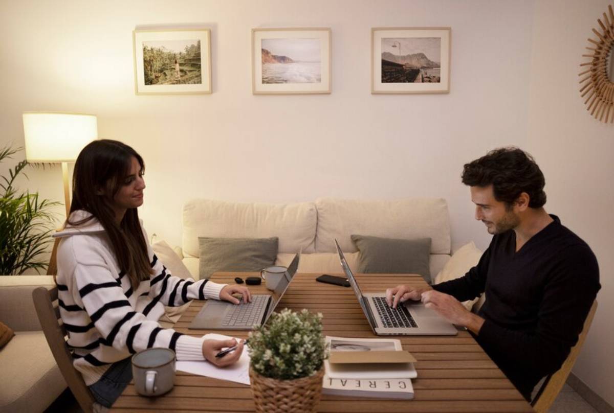 Two individuals work at a wooden table with laptops, surrounded by indoor plants and framed landscape photographs on the wall.